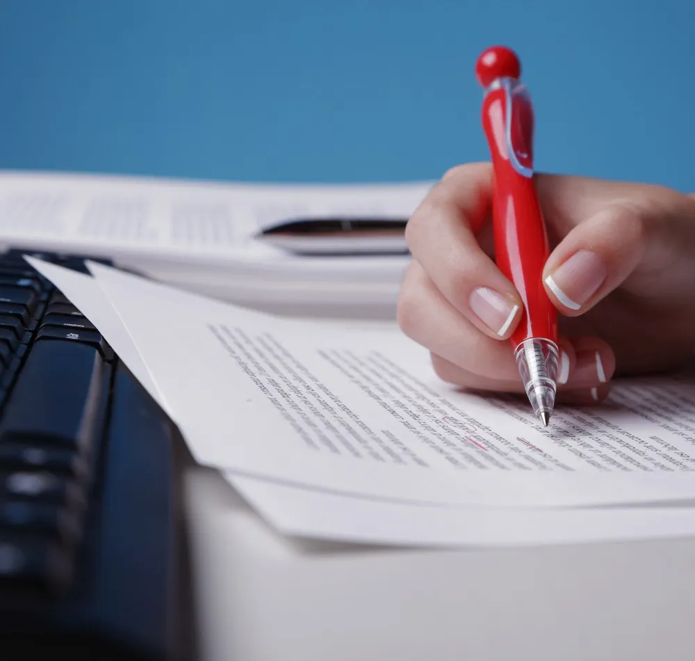 Hand with a red pen reviewing printed documents beside a keyboard