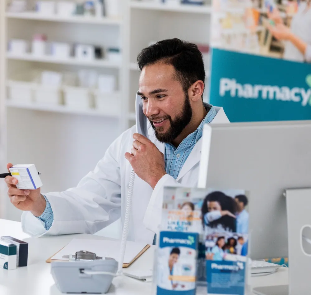 Smiling pharmacist on the phone, holding a box of medication at the pharmacy counter