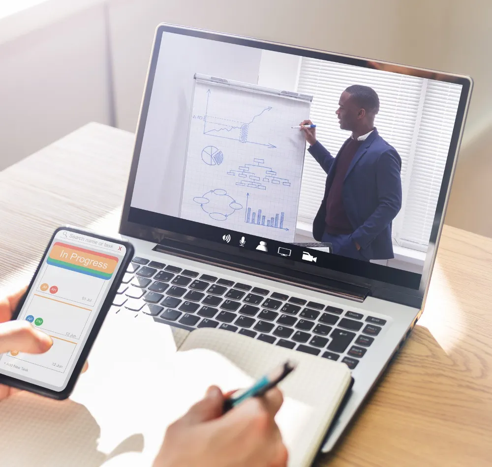 Laptop showing a presenter at a flip chart on a video call, with a hand holding a phone in front