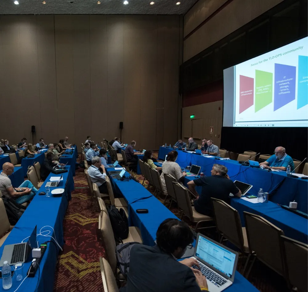 Delegates with laptops at long tables in a meeting room, slides projected on a screen
