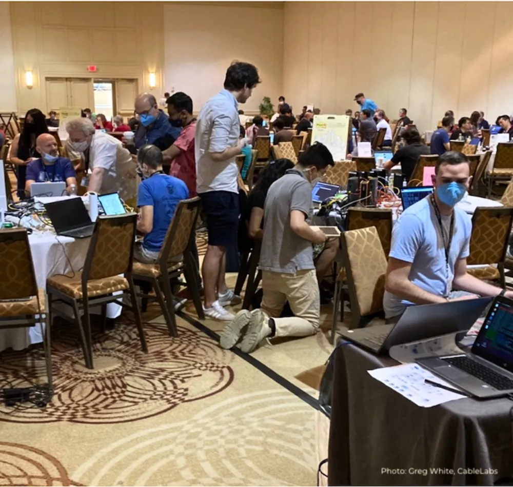 Hackathon participants, many wearing masks, working on laptops in a hotel ballroom