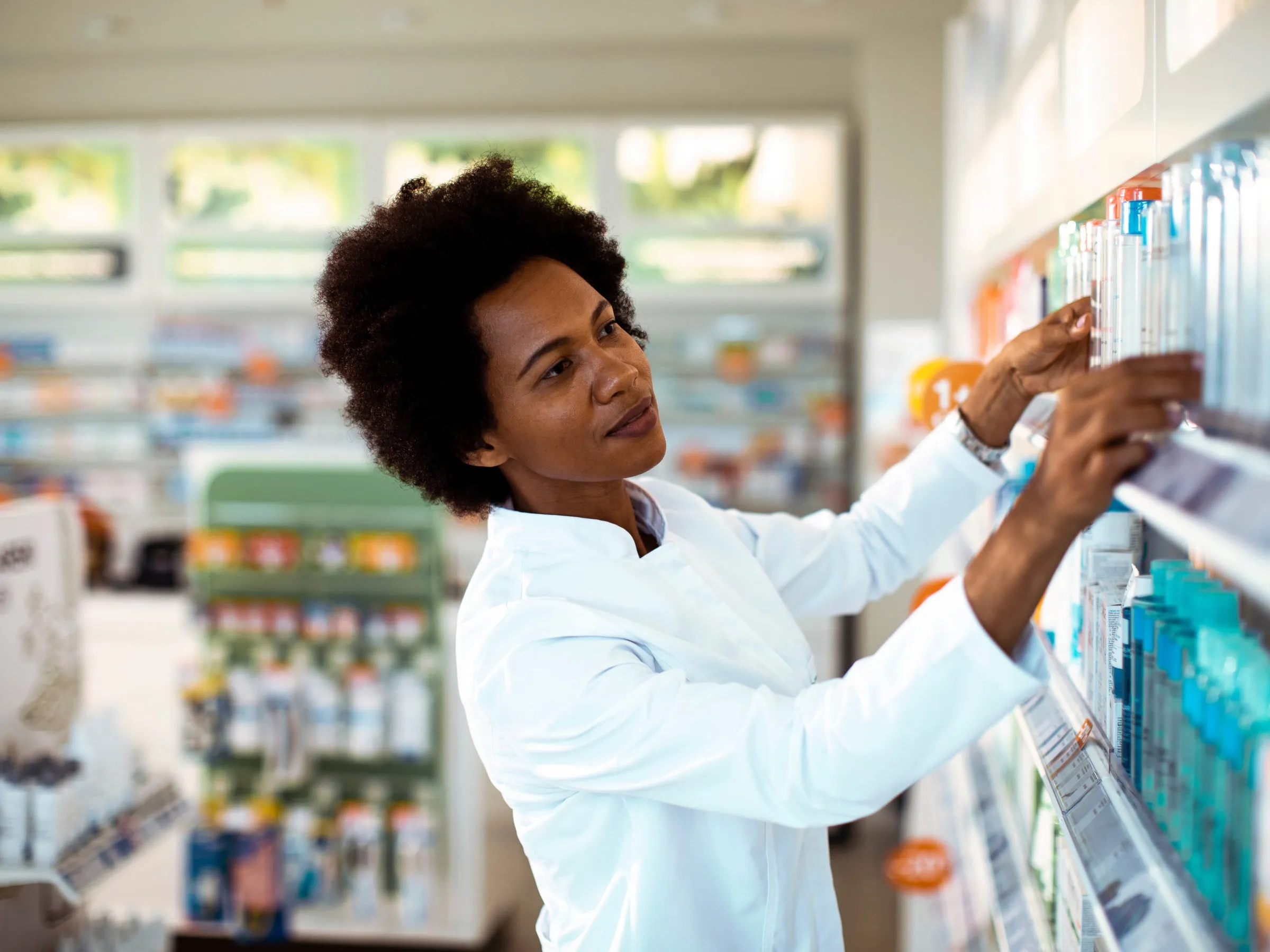 Pharmacist in a white coat reaching for a product on a pharmacy shelf